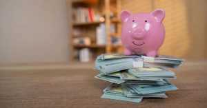 Pink piggy bank sitting on stacks of cash on a wooden table. 