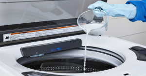 Person wearing blue gloves pouring liquid detergent from a measuring cup into a washing machine.