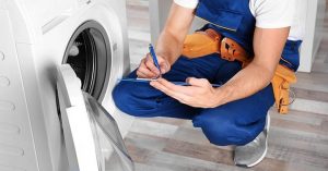 Maintenance technician in blue uniform and tool belt inspecting a washing machine and writing notes on a clipboard.