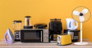 A variety of small kitchen appliances including a microwave, coffee maker, blender, toaster, electric kettle, fan, and iron placed on a wooden countertop against a yellow background.