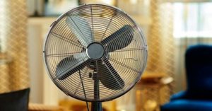 Black stand fan in a cozy, warmly lit living room with a patterned curtain in the background.