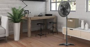 Black stand fan in a modern, cozy home office with wooden furniture, potted plant, and bright natural light from the windows.