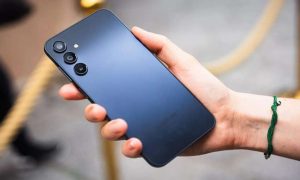 Hand holding a dark blue Samsung smartphone with a triple rear camera setup, wearing a green bracelet.
