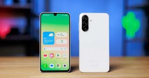 White budget smartphone with a waterdrop notch display and triple rear cameras, placed on a wooden desk.
