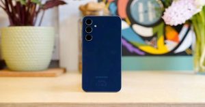 Rear view of a blue Samsung smartphone with triple camera setup, placed on a wooden desk with plants and colorful artwork in the background.
