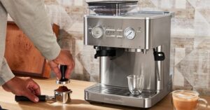 Person tamping coffee grounds next to a stainless steel espresso machine on kitchen counter.