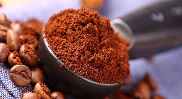 Ground coffee in a black scoop with coffee beans on fabric background.