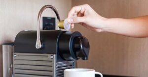 Hand inserting coffee capsule into compact espresso machine above a mug.