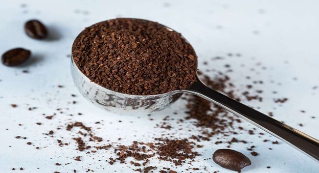 Ground coffee in metal scoop with scattered beans on white background