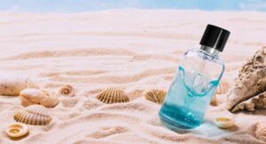 Perfume bottle with blue liquid placed on beach sand surrounded by seashells. 