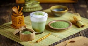 Glass of matcha latte with bamboo whisk and powdered matcha on bamboo mat