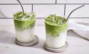 Iced matcha latte with milk in two glasses and metal straws on white coasters.