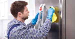Man cleaning stainless steel refrigerator door with spray and cloth.