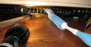 Cleaning refrigerator coils under appliance with brush and vacuum.