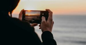 Person holding a smartphone horizontally, photographing a sunset over the ocean.