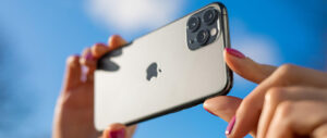 Close-up of a hand holding a silver iPhone with triple cameras against blue sky.