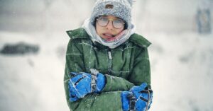 A young boy bundled in a green winter coat and hat, shivering in the snow.