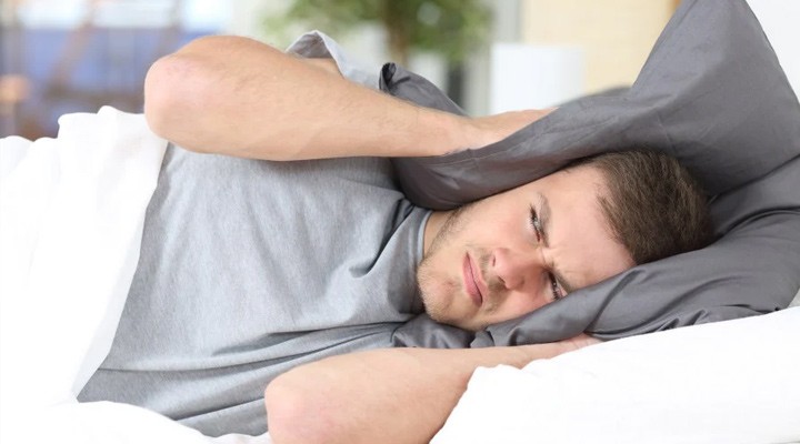 Man in bed covering his ears with a gray pillow, disturbed by loud noise.