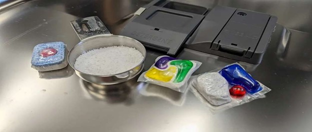 Different dishwasher detergents including powder, liquid gel, tablets, and detergent pods placed next to the dishwasher dispenser