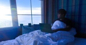 Man using laptop in bed near window with morning sunlight and coffee cups 