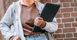 Person holding closed laptop while standing outdoors near brick wall 