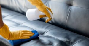 Cleaning a black leather sofa with spray and microfiber cloth.