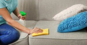 Cleaning a beige fabric sofa with spray bottle and yellow cloth.