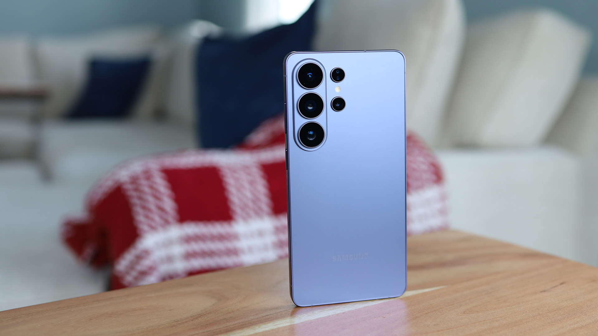 Samsung Galaxy S26 Ultra in light blue color standing on a wooden table with a blurred indoor background. 