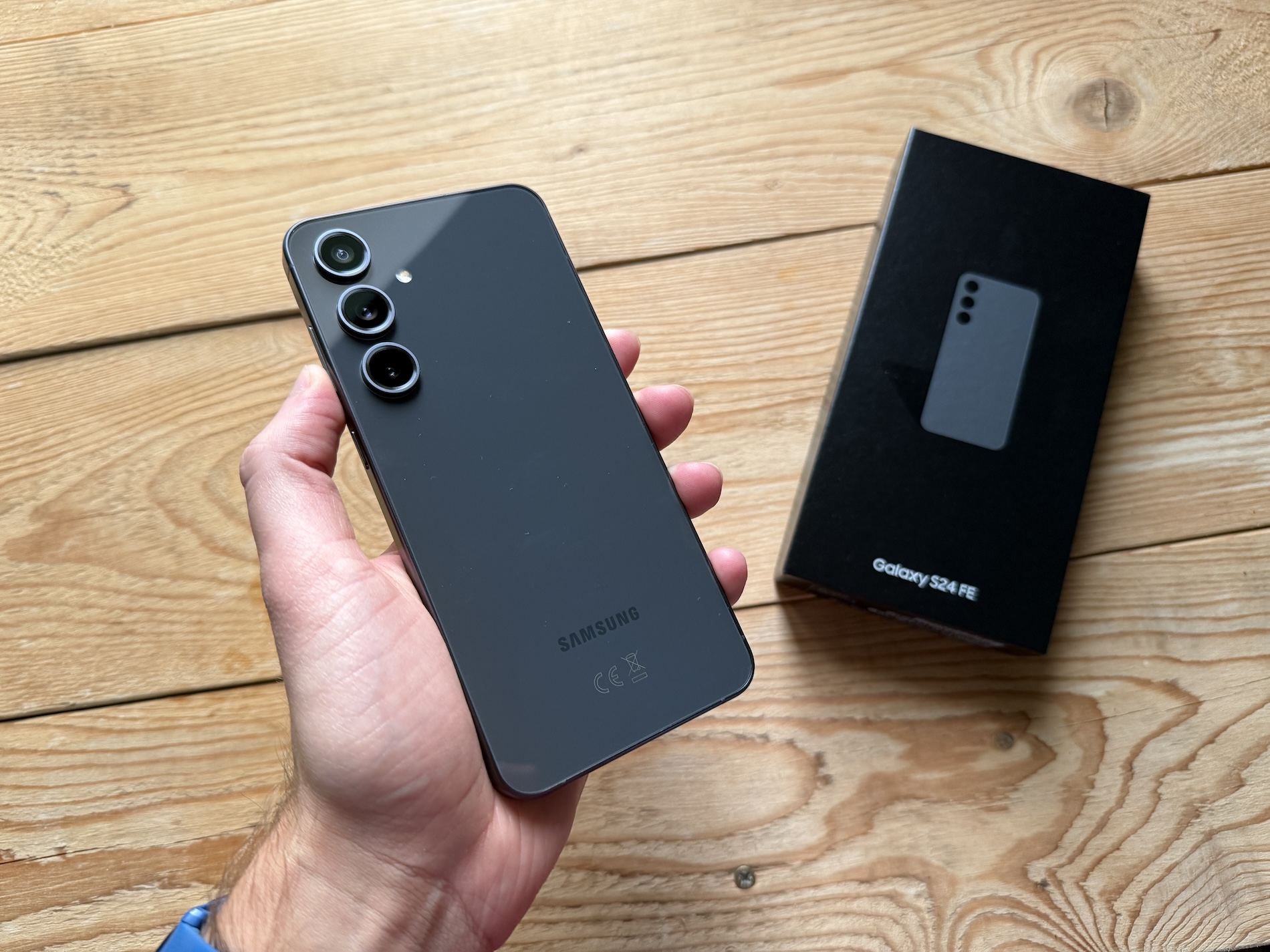 Samsung Galaxy S24 FE smartphone in graphite color held in hand, showcasing the rear triple-camera system with the original retail box on a wooden surface.