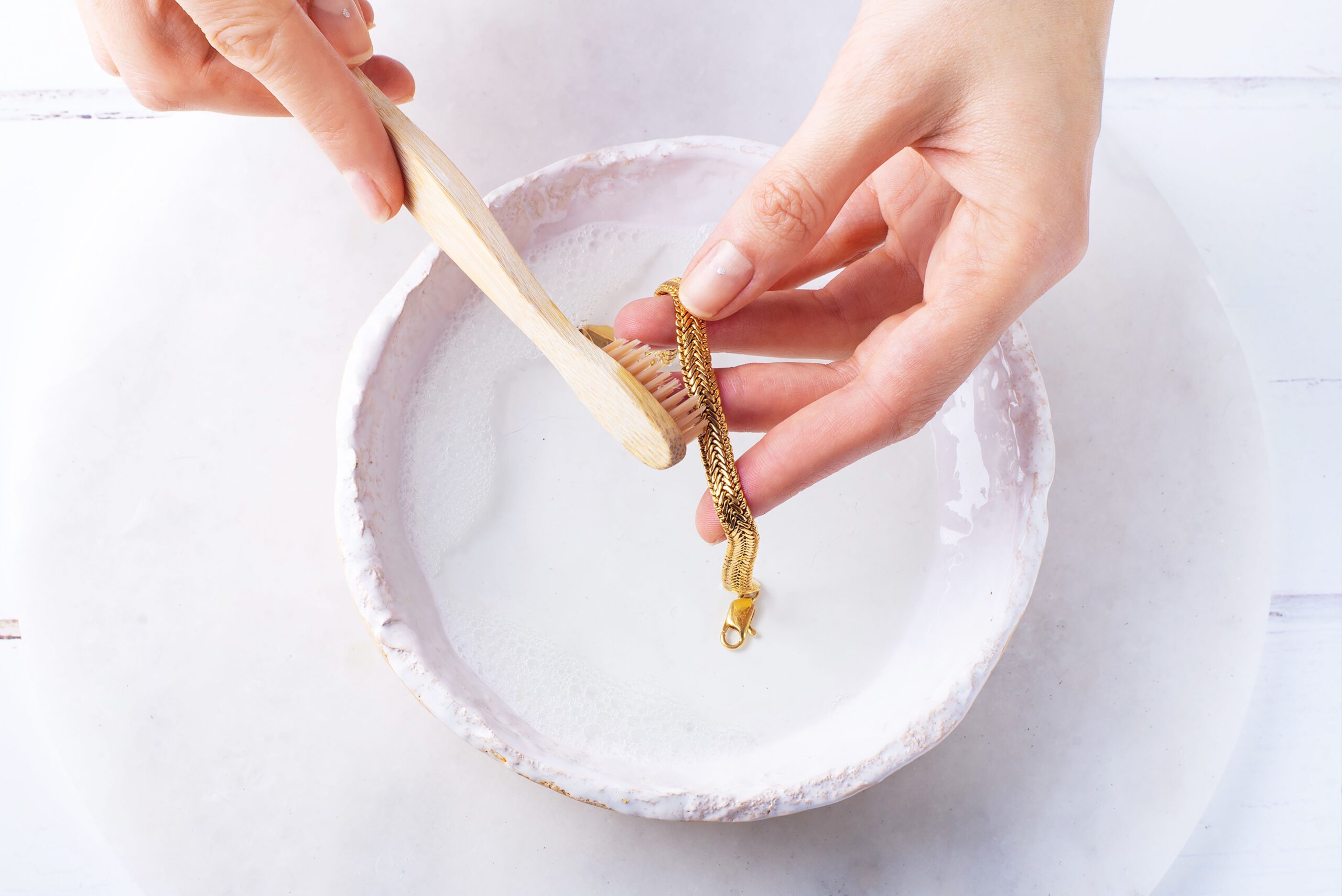 Person cleaning a gold bracelet with a soft brush in soapy water.
