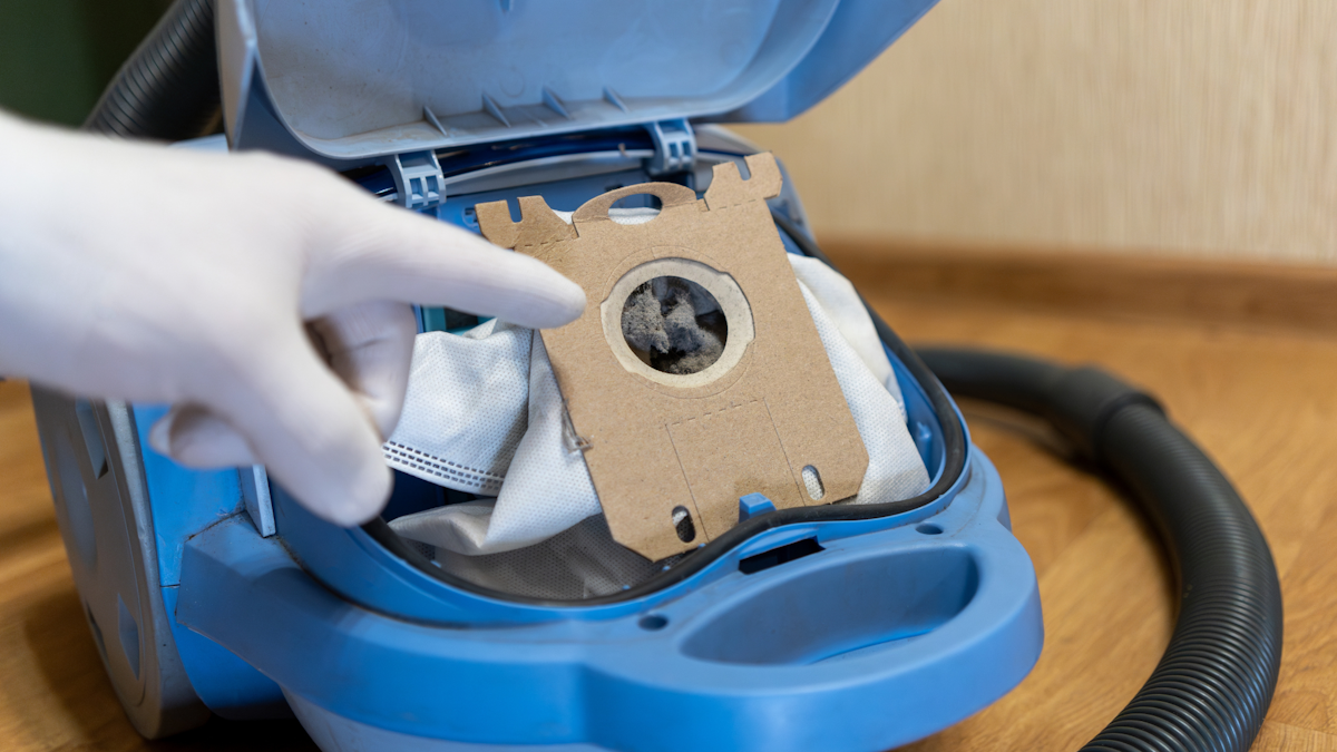 Gloved hand pointing at a full dust bag inside an open vacuum cleaner
