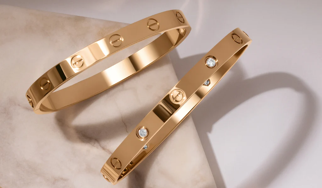 Two polished gold bangles with screw details and embedded diamonds displayed on a marble surface.
