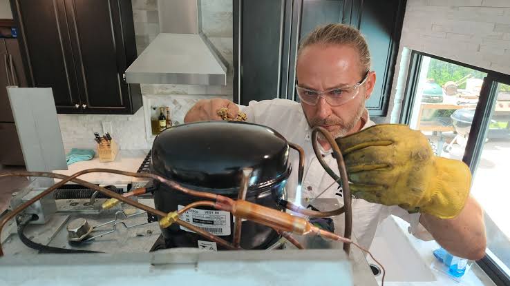 Technician inspecting a refrigerator compressor and copper tubing while wearing protective gloves and glasses.