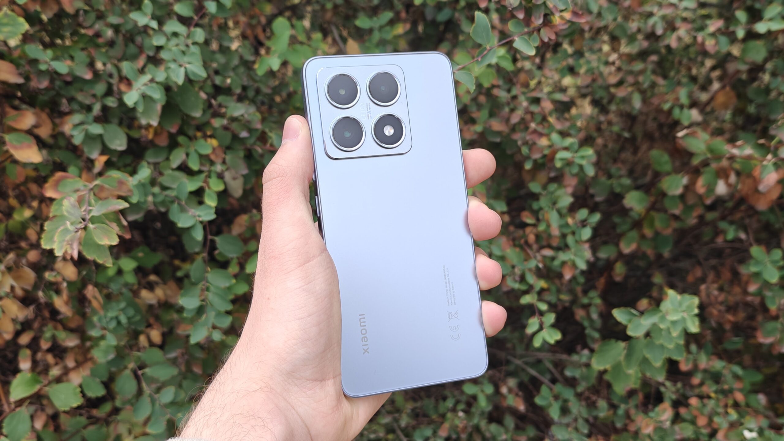 Hand holding a silver Xiaomi smartphone outdoors against green foliage.