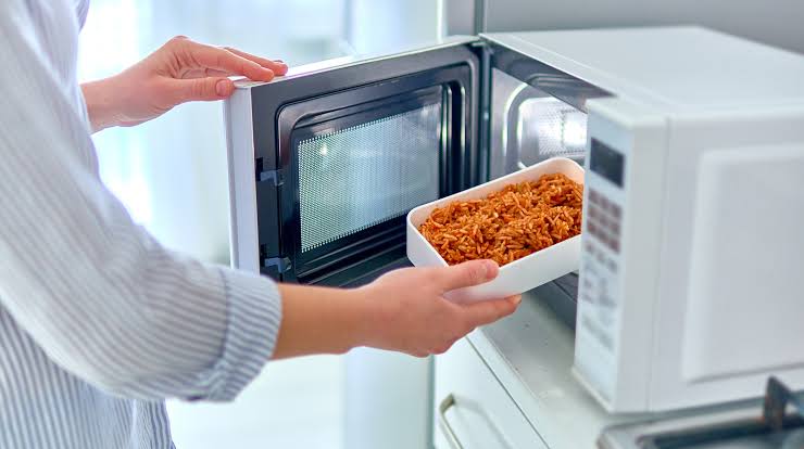 Person placing a dish of cooked food inside an open microwave oven.