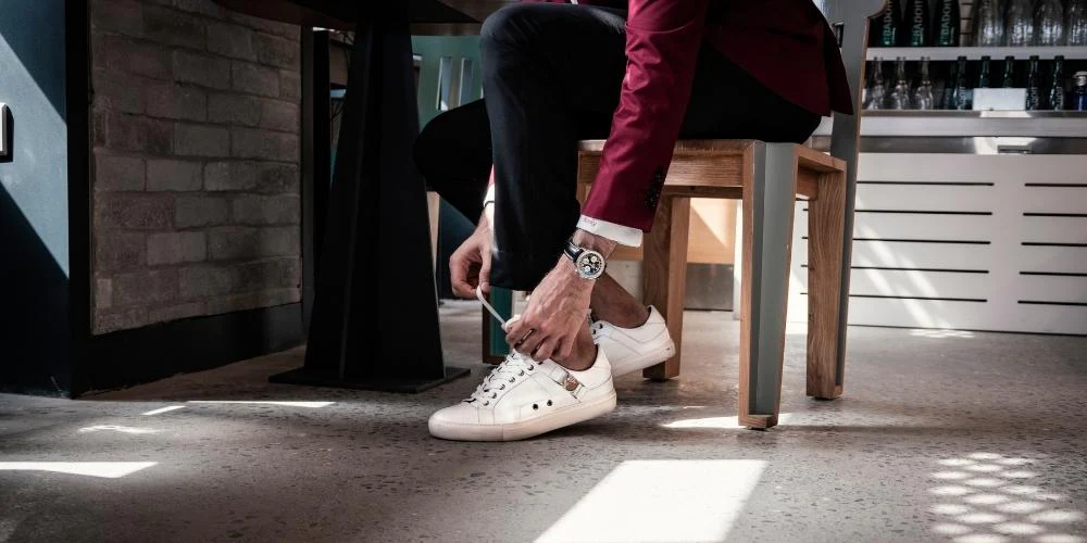 Person wearing white sneakers while sitting and tying shoelaces indoors.
