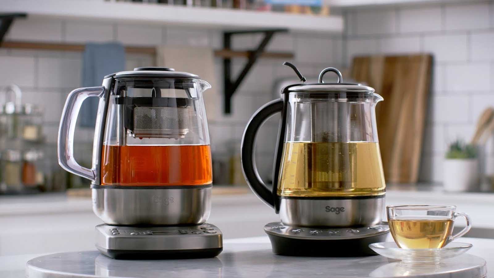 Two glass electric tea makers with tea infusers brewing black and green tea on a kitchen counter.