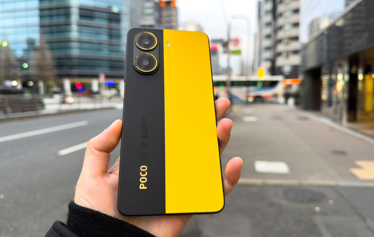 Hand holding a yellow and black Poco smartphone on a blurred city street.