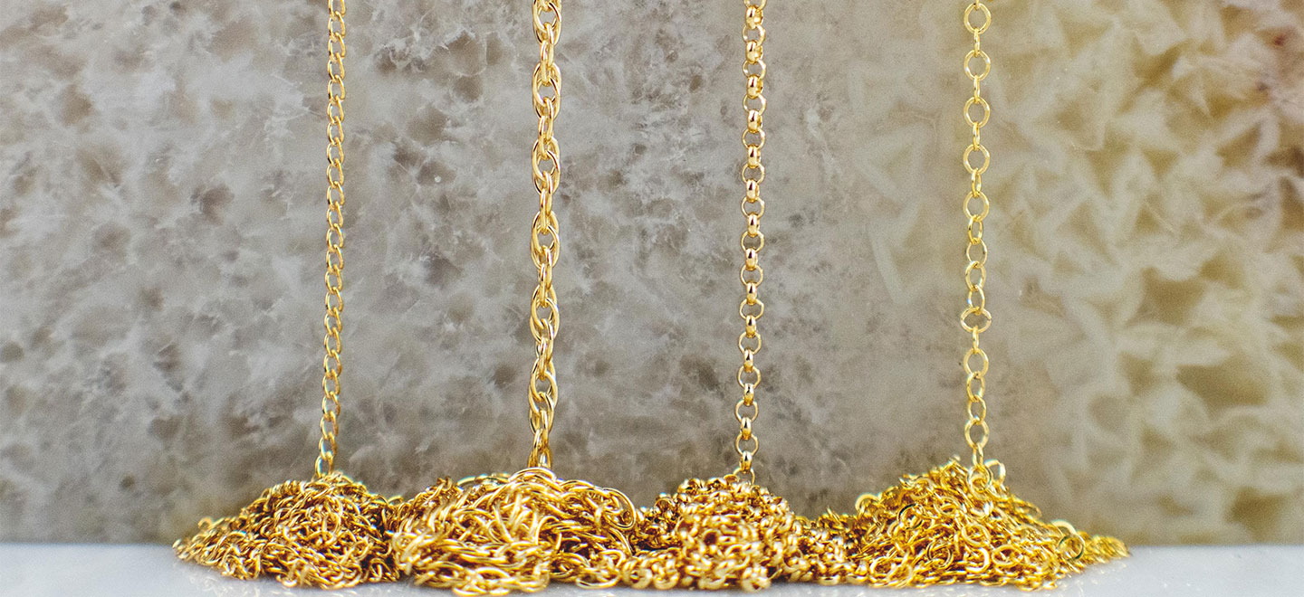Different styles of gold chains hanging above tangled piles on a stone surface.
