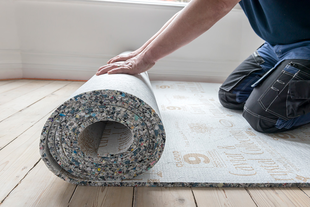 Person installing carpet padding under a rug on a wooden floor.