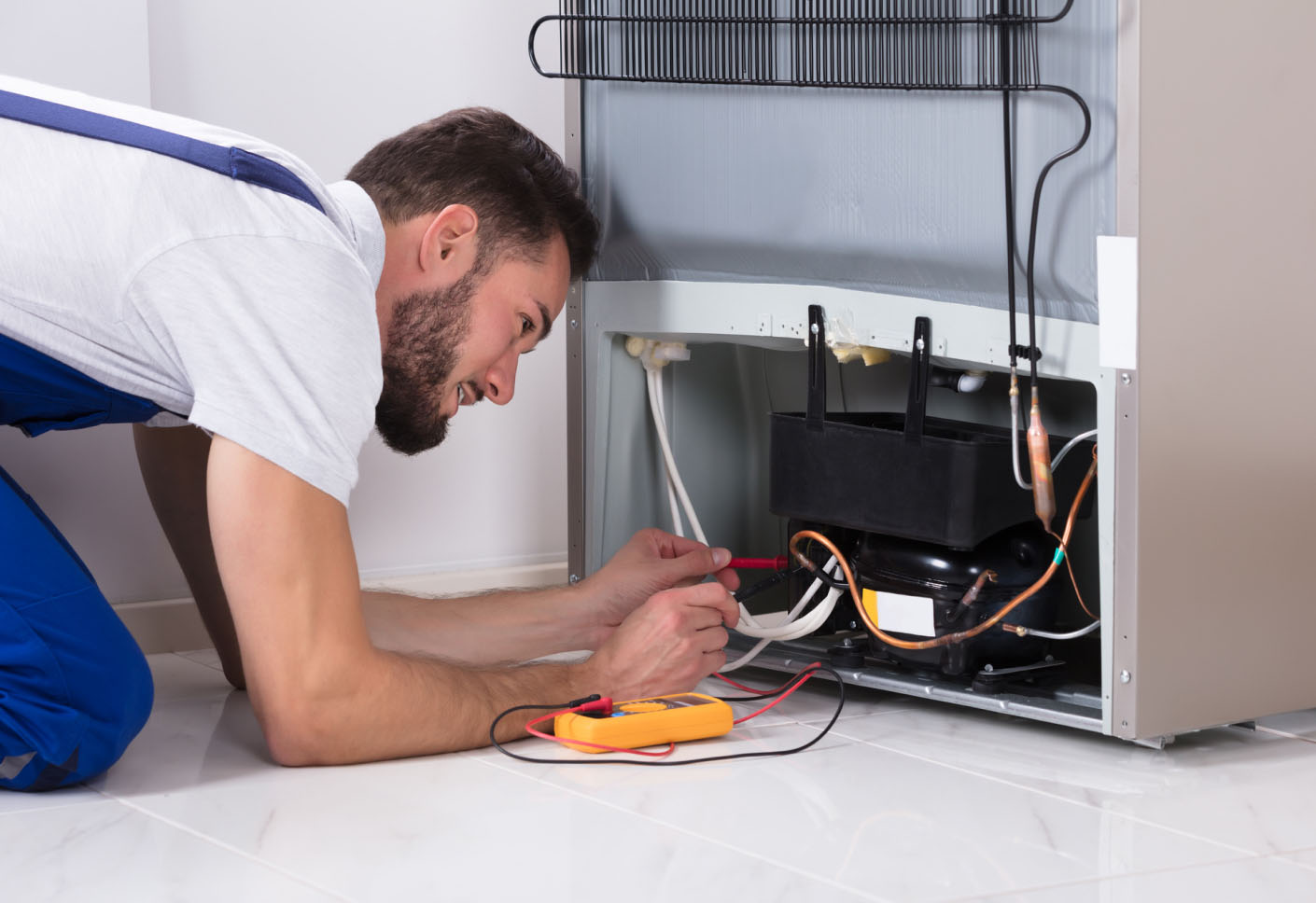 Technician inspecting and testing a refrigerator compressor with a digital multimeter.