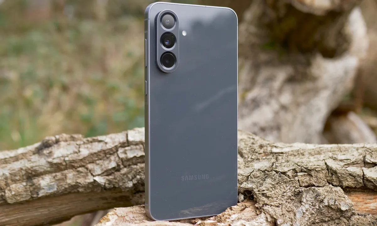 Samsung smartphone with triple rear cameras standing on a tree log outdoors.
