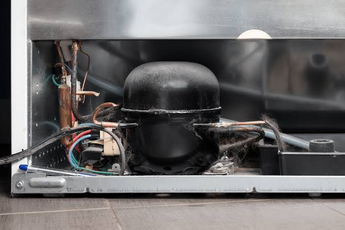 Dust-covered refrigerator compressor installed behind the appliance with visible cooling components.