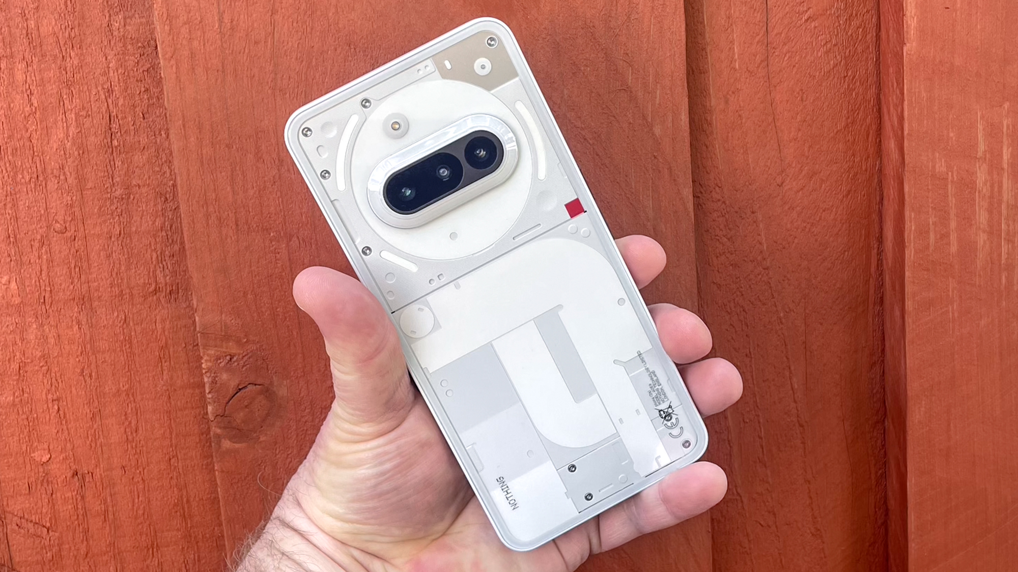 Hand holding a white Nothing Phone against a red wooden background.
