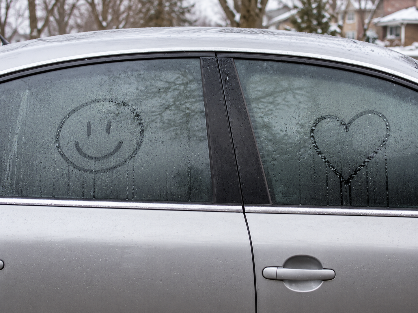 Fogged car side windows with smiley and heart drawings on a parked silver car.